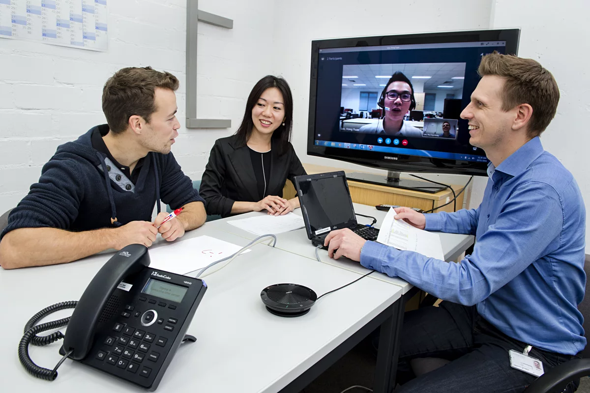 Three people sitting at a desk and talking in a hybrid video meeting.
