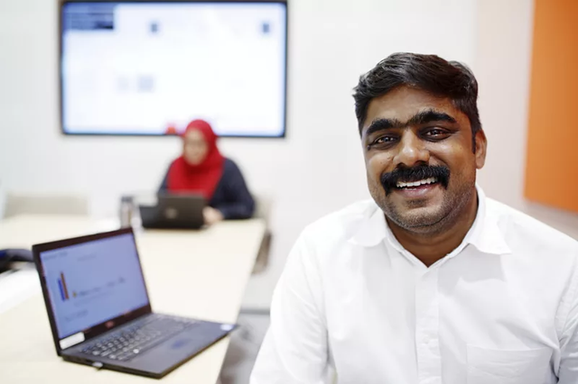 A man smiles at the camera in front of a laptop and a women sitting in the back and working on her labtop.