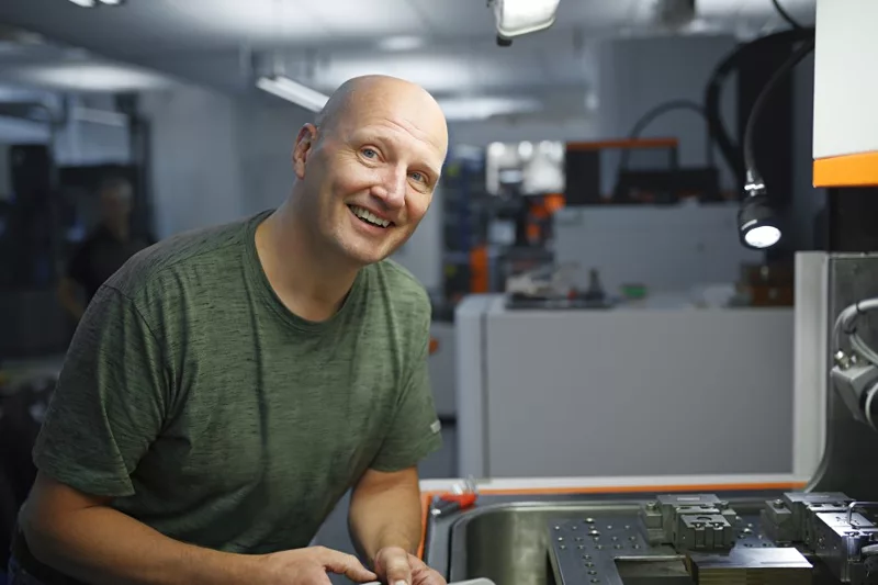 A man in a green shirt smiles while working on an industrial machine.